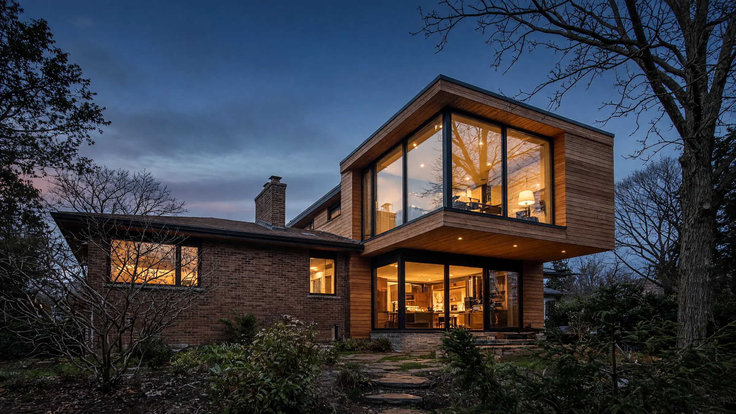 A 1960s brick bungalow at dusk with a cantilevered two-storey cedar addition, its tall windows glowing warm against a deep blue sky